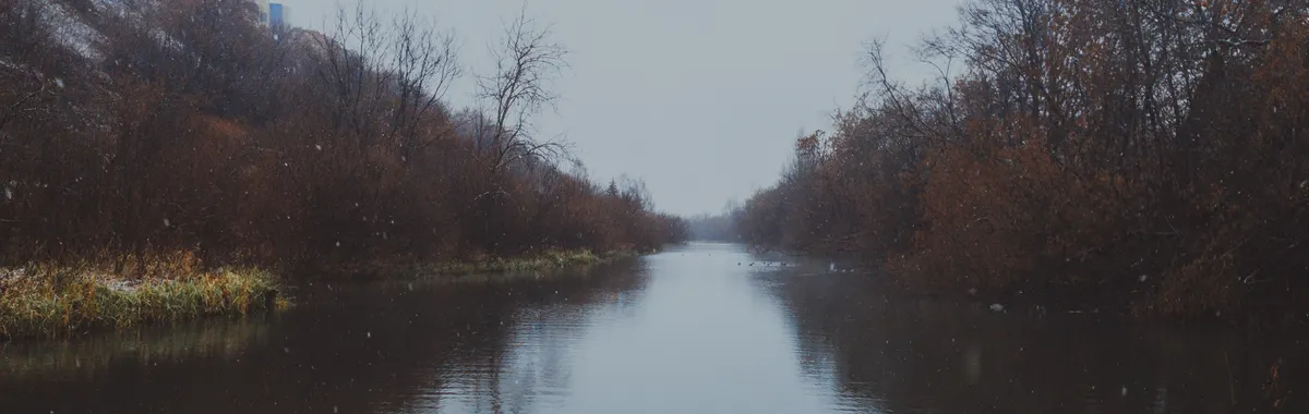 A serene view of a river in winter, surrounded by trees and hills, under snowfall in Kirov, Russia.