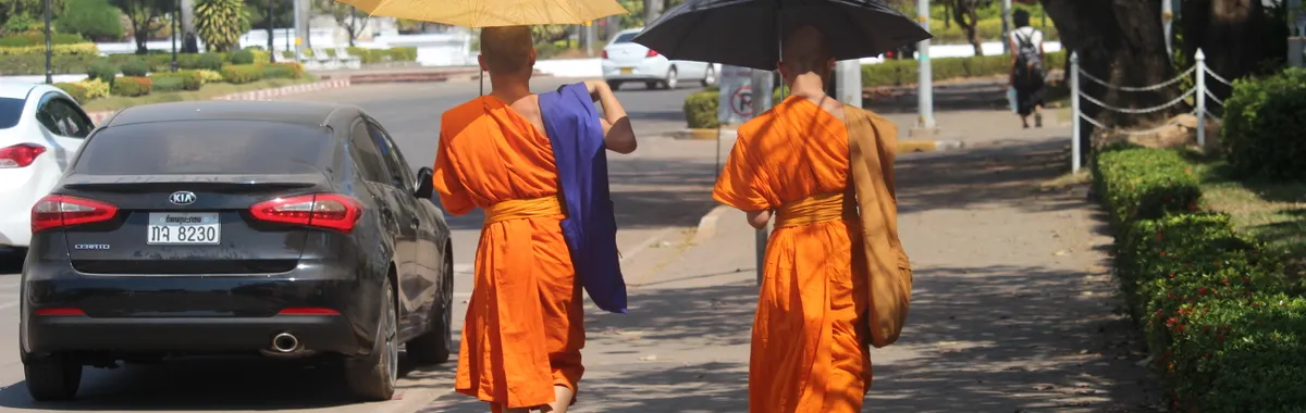Two Buddhist monks walking on a sunny sidewalk in Vientiane, Laos with umbrellas for shade.