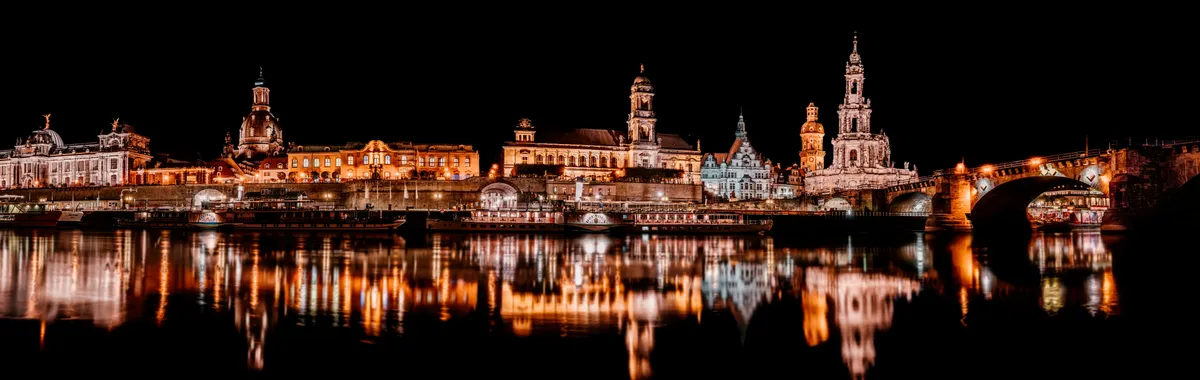 Nighttime view of illuminated Dresden buildings reflecting in the river, showcasing urban architecture.
