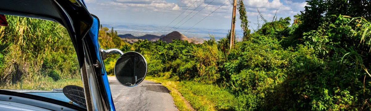 קובה: A vintage car drives along a lush rural road under a vibrant blue sky with scattered clouds.