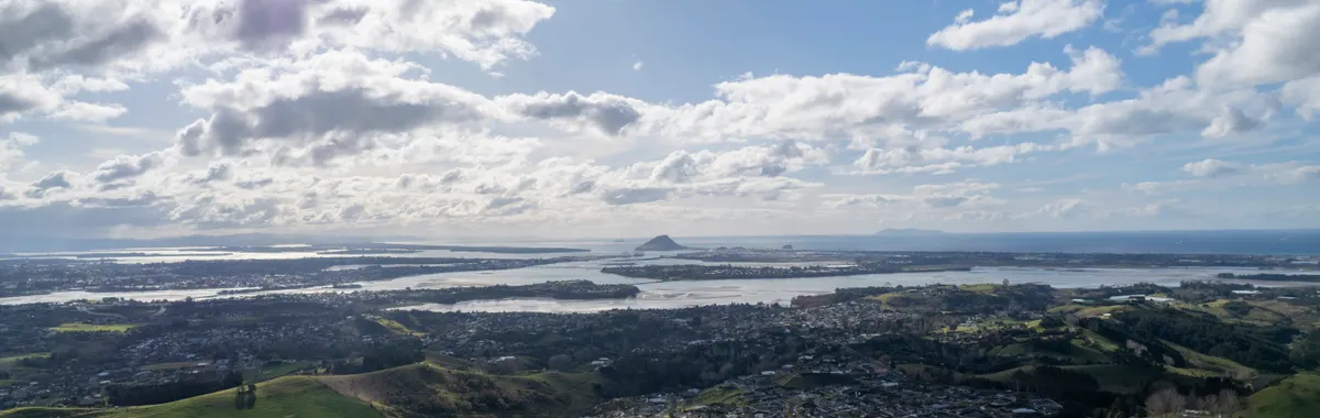 Stunning aerial view of Tauranga, New Zealand with hills, coastline, and clouds under a bright sky.