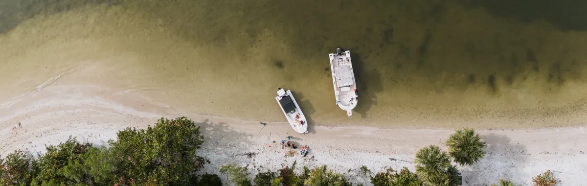 Drone shot of boats moored on the sandy shore of Lakewood Park, FL, with lush trees and clear waters.