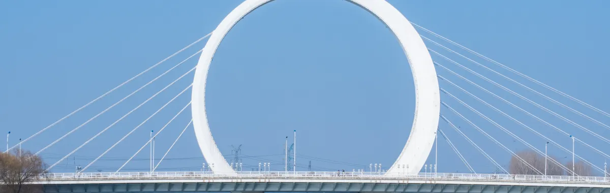 The iconic Infinity Ring Bridge in Zhengzhou, China, spanning over a tranquil river under a clear sky.