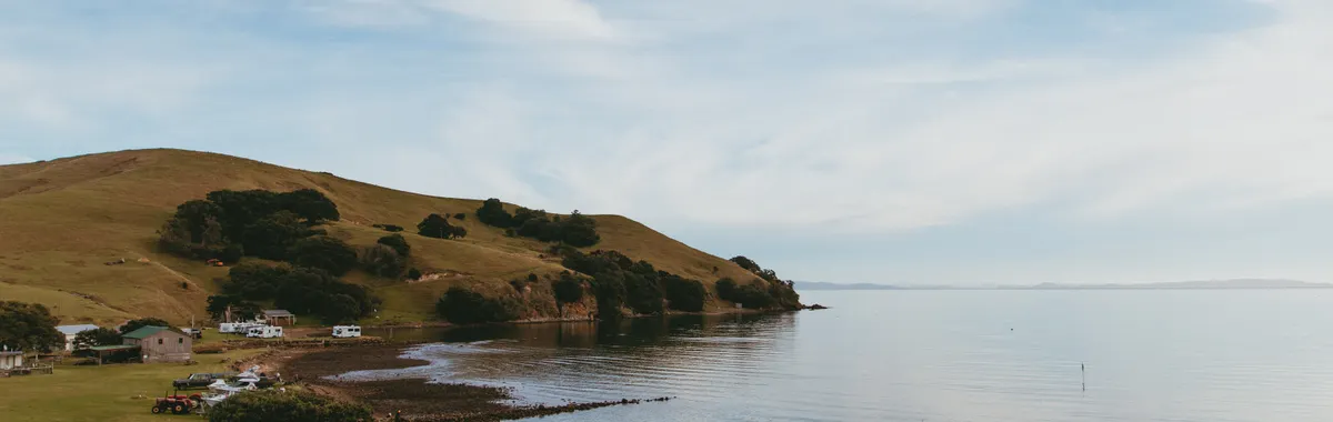 ניו זילנד: Tranquil coastal landscape at Kereta, Waikato, New Zealand. Rolling hills meet the serene ocean shore under a blue sky.
