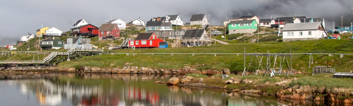 גרינלנד: Picturesque Qeqertarsuaq village reflecting on a tranquil lake in Greenland under a cloudy sky.