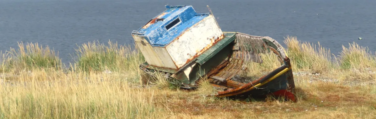 Worn-out boat resting on grassy shore with ocean and mountain backdrop in Punta Arenas, Chile.