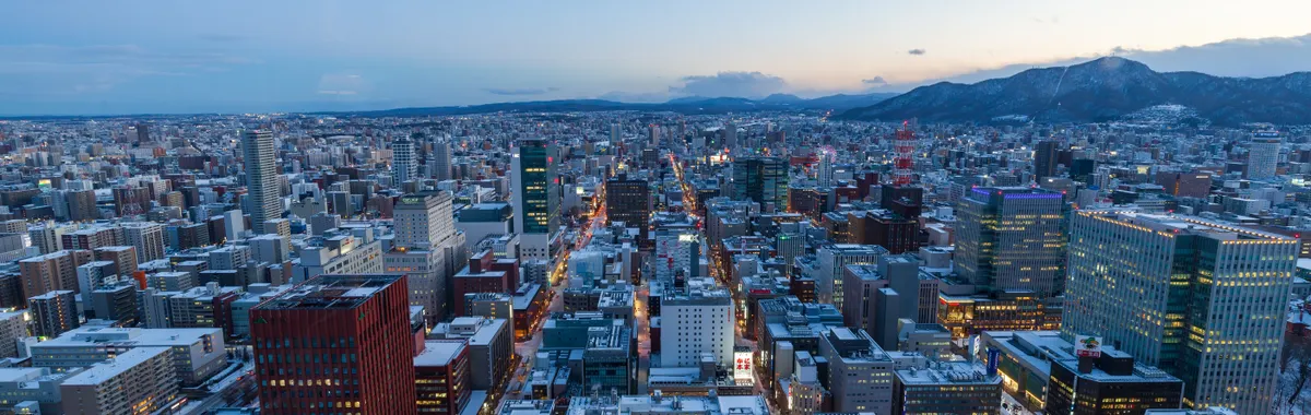 Captivating aerial view of Sapporo, Japan in winter at dusk with snow-covered buildings and distant mountains.
