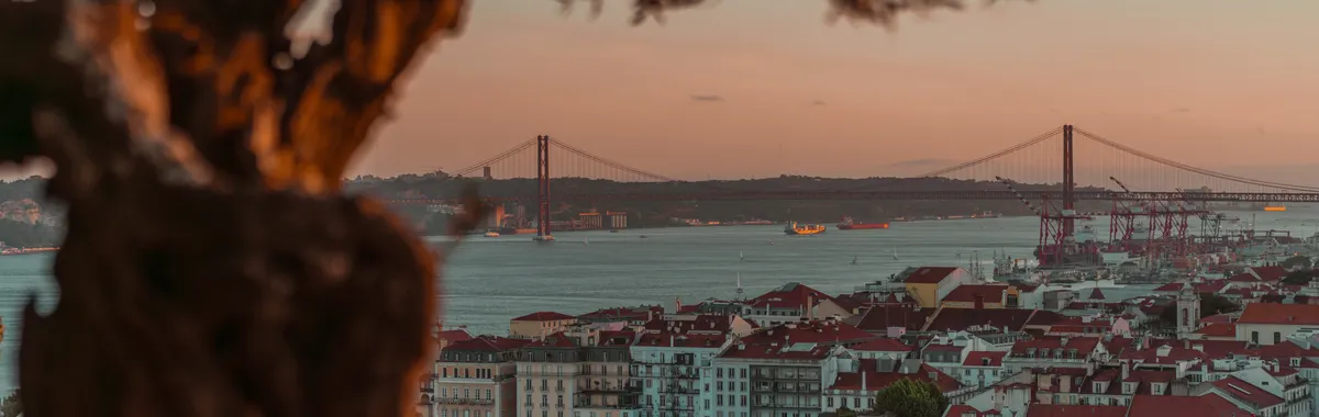 Beautiful sunset over Lisbon with a view of the iconic 25 de Abril Bridge, framed by trees.