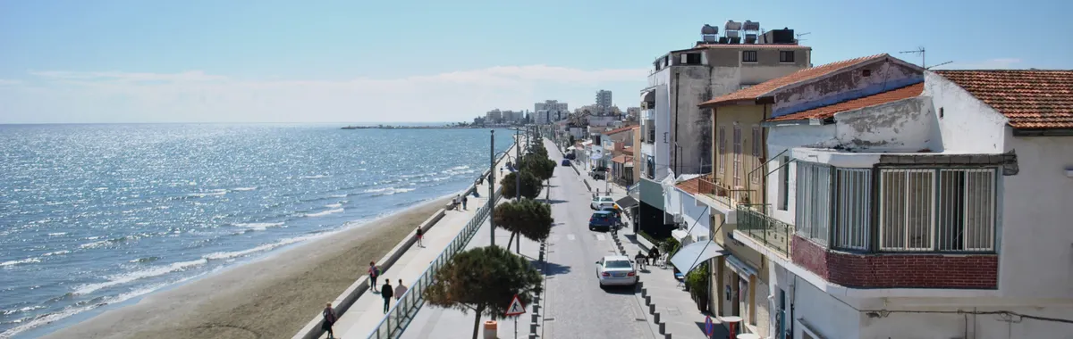 A scenic street along the coast in Larnaca, Cyprus with clear blue skies, waterfront, and walking pedestrians.