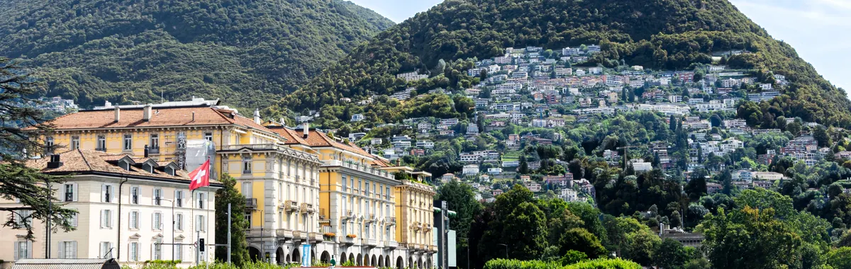 Scenic view of Lake Lugano with mountains and elegant buildings against a bright sky.