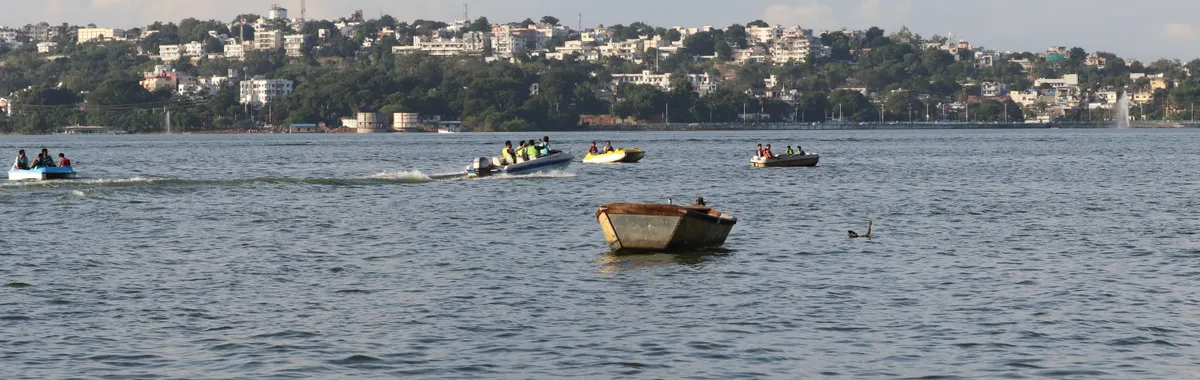Boats float on Upper Lake in Bhopal with a cityscape in the background under a sunny sky.