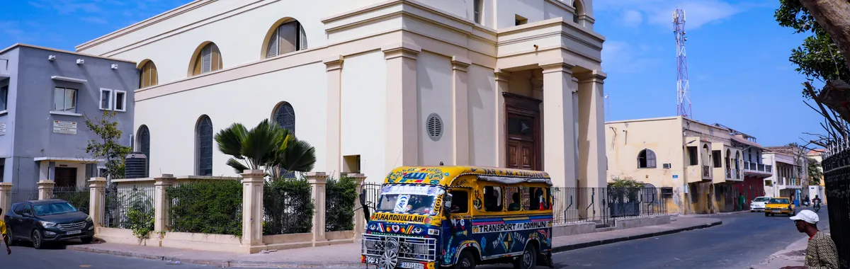 A vibrant minibus travels the streets of Dakar, passing a historic church under a clear blue sky.
