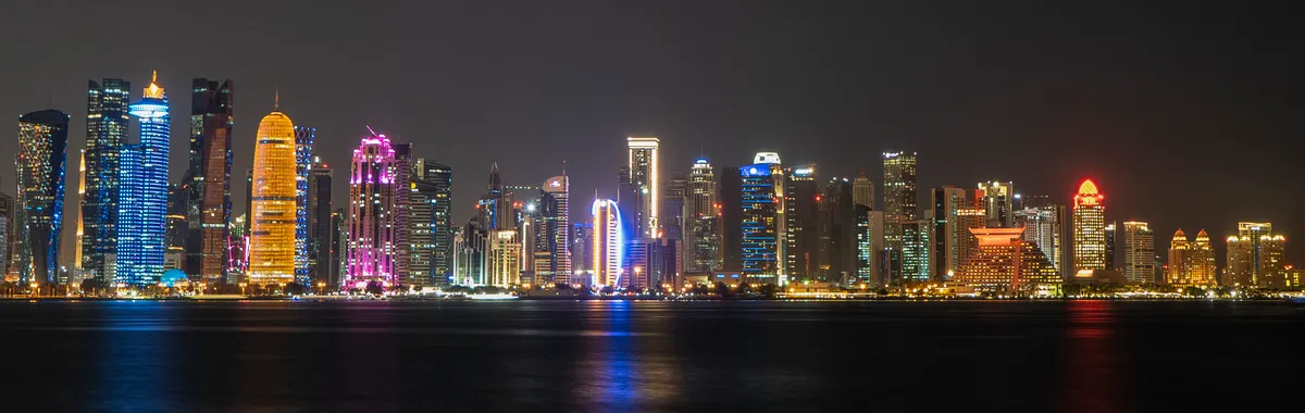 קטאר: Doha cityscape featuring illuminated skyscrapers reflecting on the water at night.
