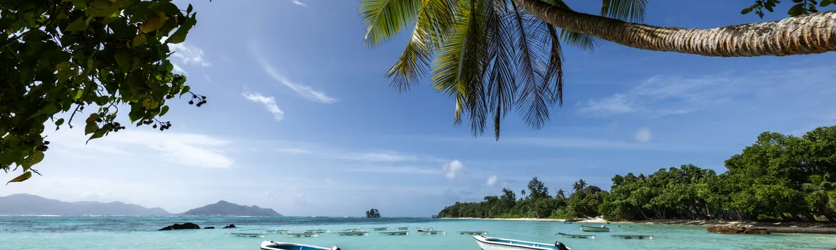 איי סיישל: Serene tropical beach in Seychelles with clear waters, boats, and lush palm trees.