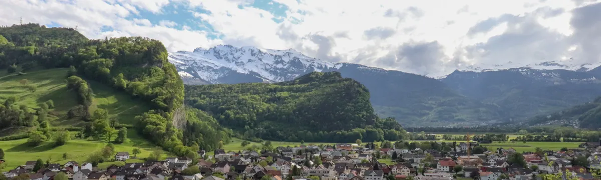 ליכטנשטיין: Picturesque Liechtenstein village with snowcapped mountains and lush hills.