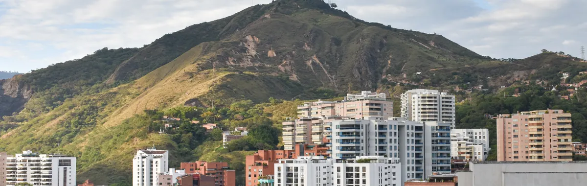 A vibrant cityscape of Cali, Colombia, with lush Andes mountains and modern buildings under a cloudy sky.