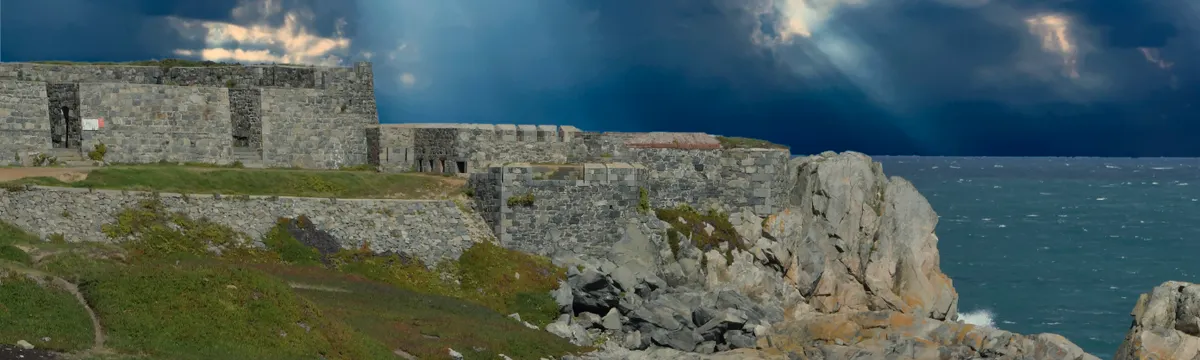 גרנזי: Rugged fort along Guernsey's rocky coast with dramatic clouds and sunrays breaking through.