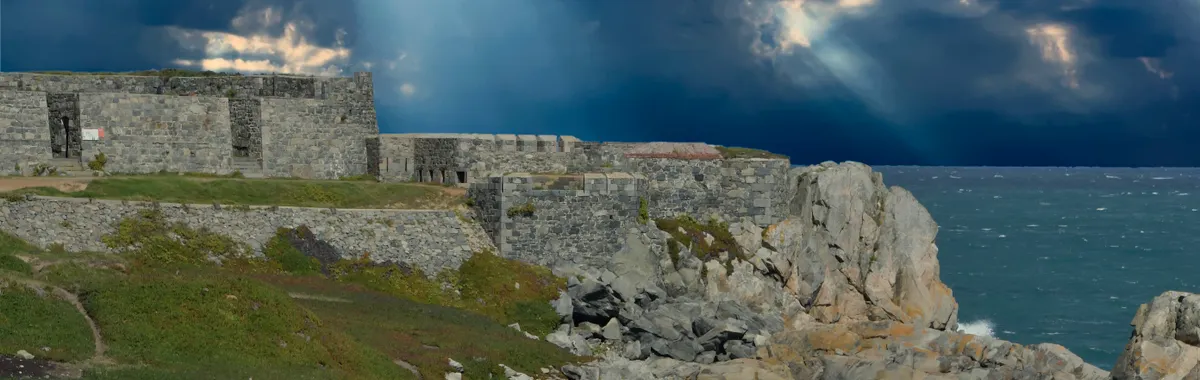 גרנזי: Rugged fort along Guernsey's rocky coast with dramatic clouds and sunrays breaking through.