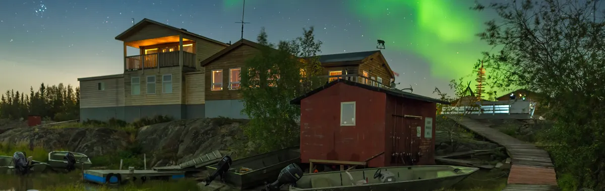 Colorful aurora borealis over rustic lakeside cabins and boats in Yellowknife, Canada.